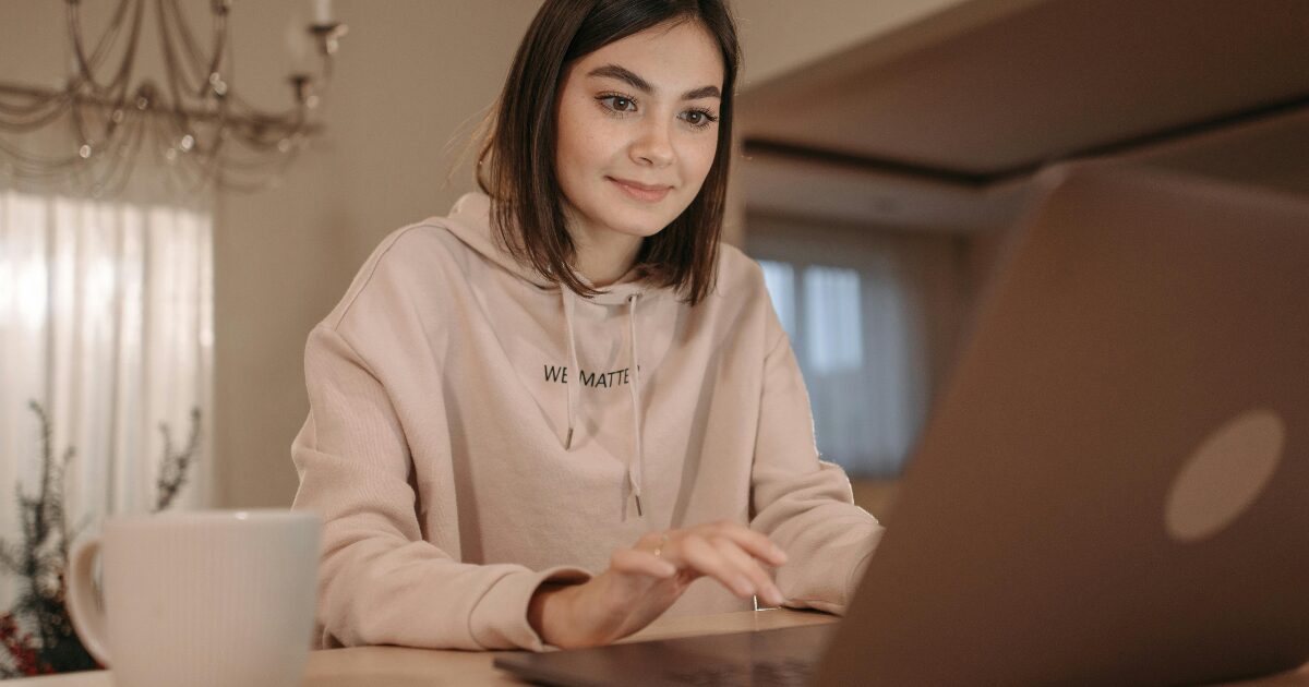 A Caucasian woman wearing a cream-colored hoodie smiling while typing on a laptop