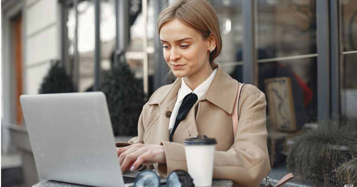 A Caucasian woman wearing a tan coat smirking while using her laptop.