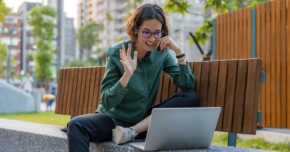 A Caucasian woman wearing a teal buttoned shirt smiling and waving in front of a laptop
