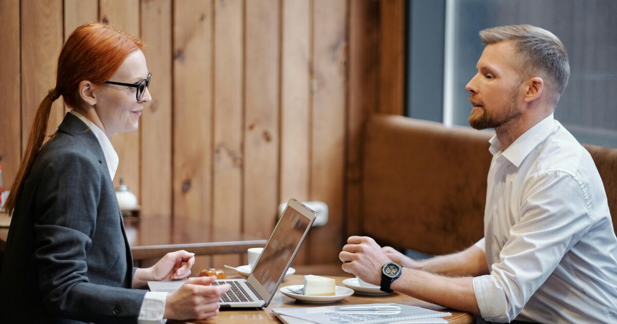 A Caucasian woman wearing a black blazer and a Caucasian man wearing a white buttoned shirt sitting face-to-face at a table, engaged in conversation.