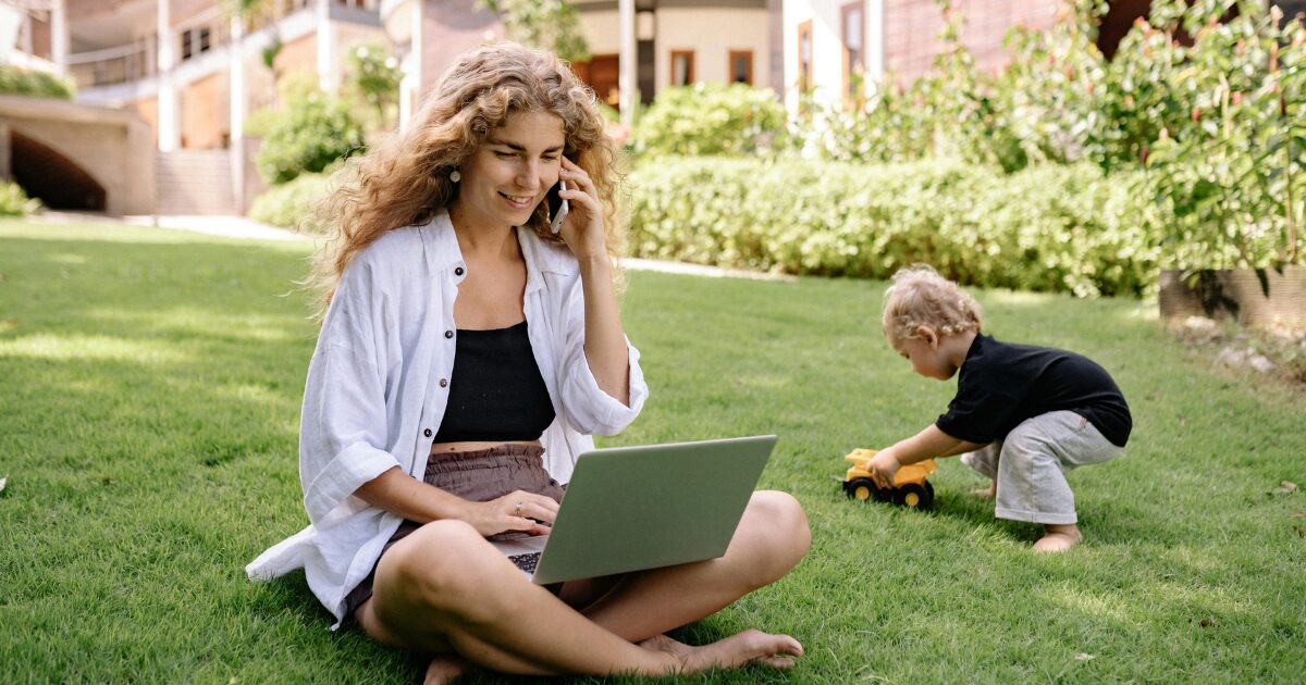 A Caucasian woman wearing a white shirt sits cross-legged on the grass with a laptop on her lap, holding a phone to her left ear. A young Caucasian child plays in the background.