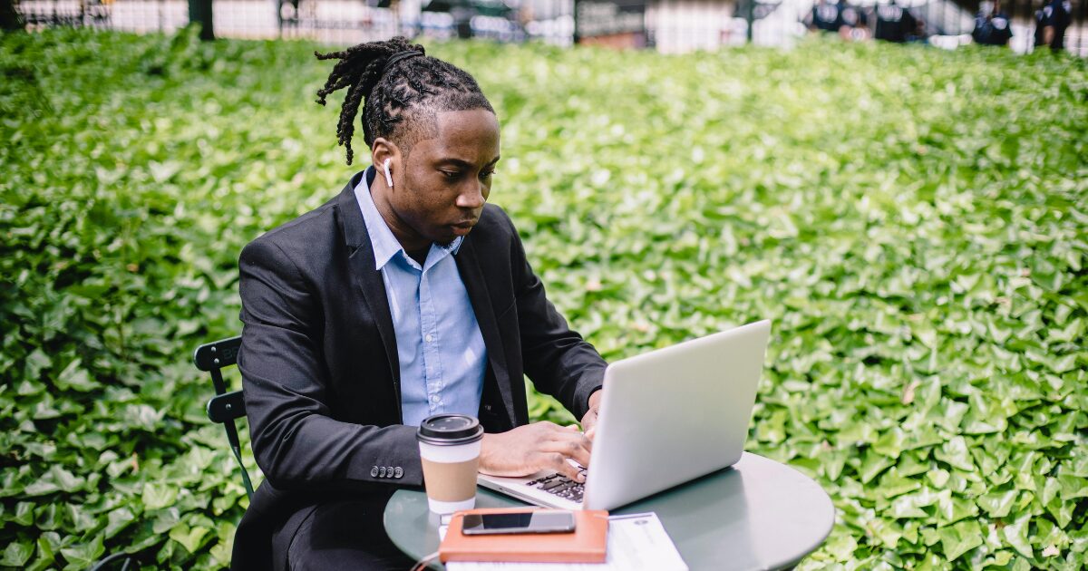 348 A Black man wearing a black blazer sitting outdoors while using his laptop.