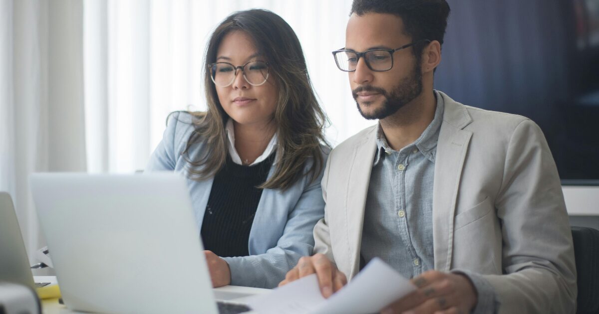 349 An East Asian woman in a pastel blue blazer and a South Asian man in a light grey blazer look at a laptop together. The man holds a document in his hands.