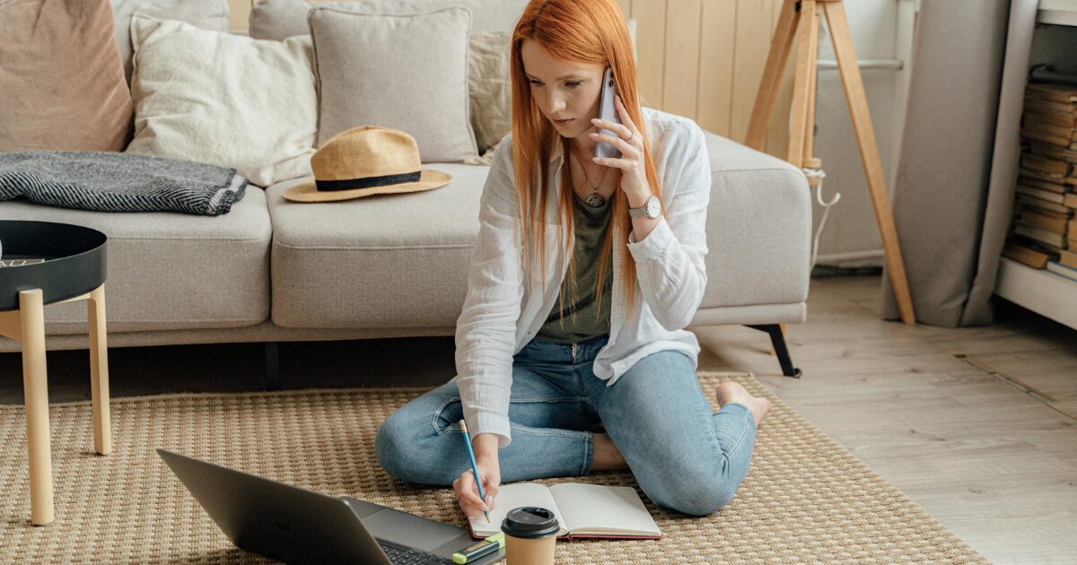 354 A Caucasian woman wearing a white blazer sits on the floor, writing in a notebook while holding a phone in her left hand.