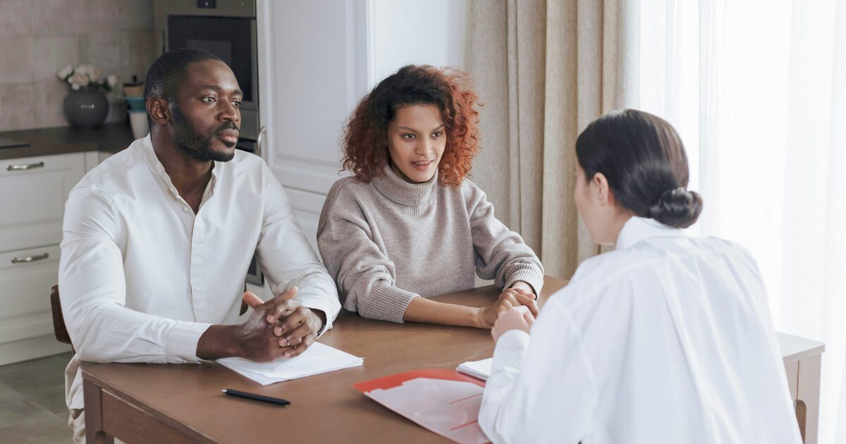 363 A Black man wearing a white shirt and a Black woman wearing a beige sweater sitting on a table across a Caucasian woman wearing a white blouse.