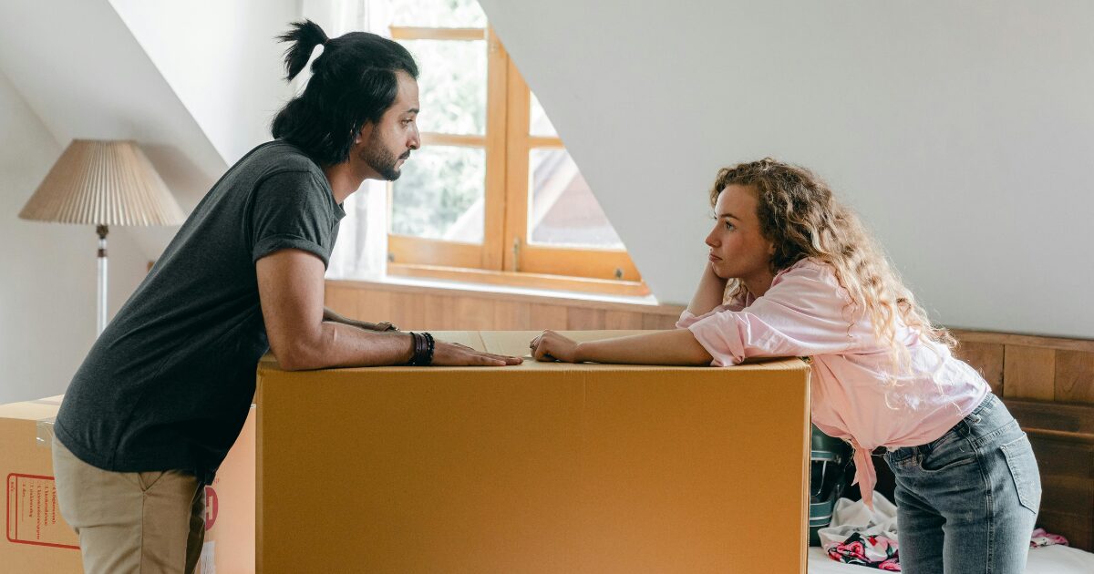 364 A Caucasian man wearing a dark grey t-shirt leans on a large box across from a Caucasian woman wearing a pink blouse, with both of them resting on the box facing each other.