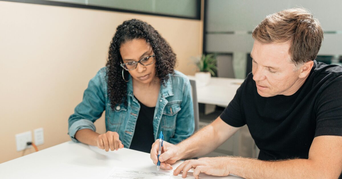 365 A Caucasian man wearing a black shirt signs a document while a Black woman wearing a denim jacket watches beside him.