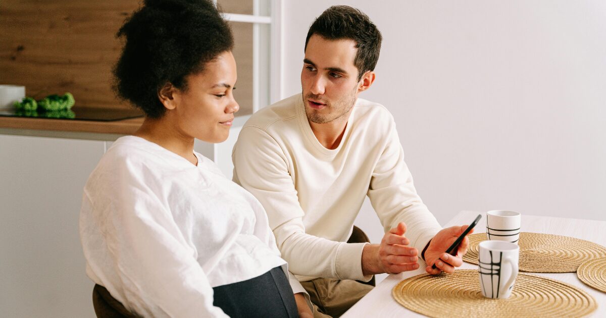 366 A Black woman wearing a white blouse sits beside a white man wearing a cream sweater. The man holds a phone and looks at the woman, while she focuses on the screen in his hands.