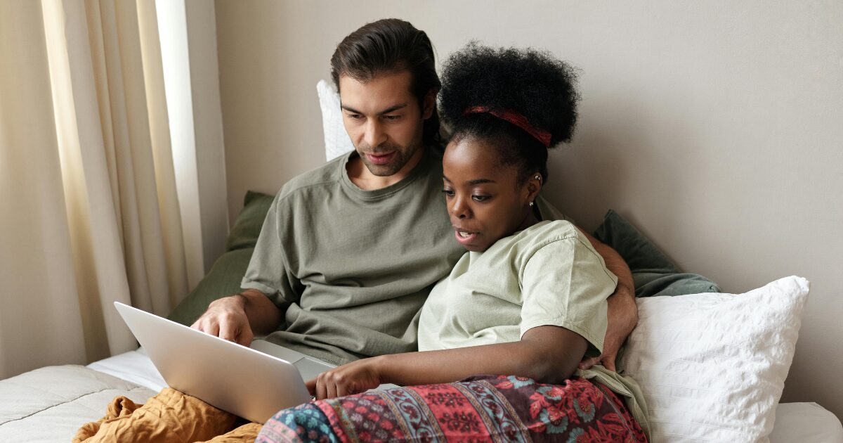 368 A Black woman wearing a light grey t-shirt cuddles with a Caucasian man wearing a green t-shirt. The man uses a laptop on his lap while they both look at the screen.