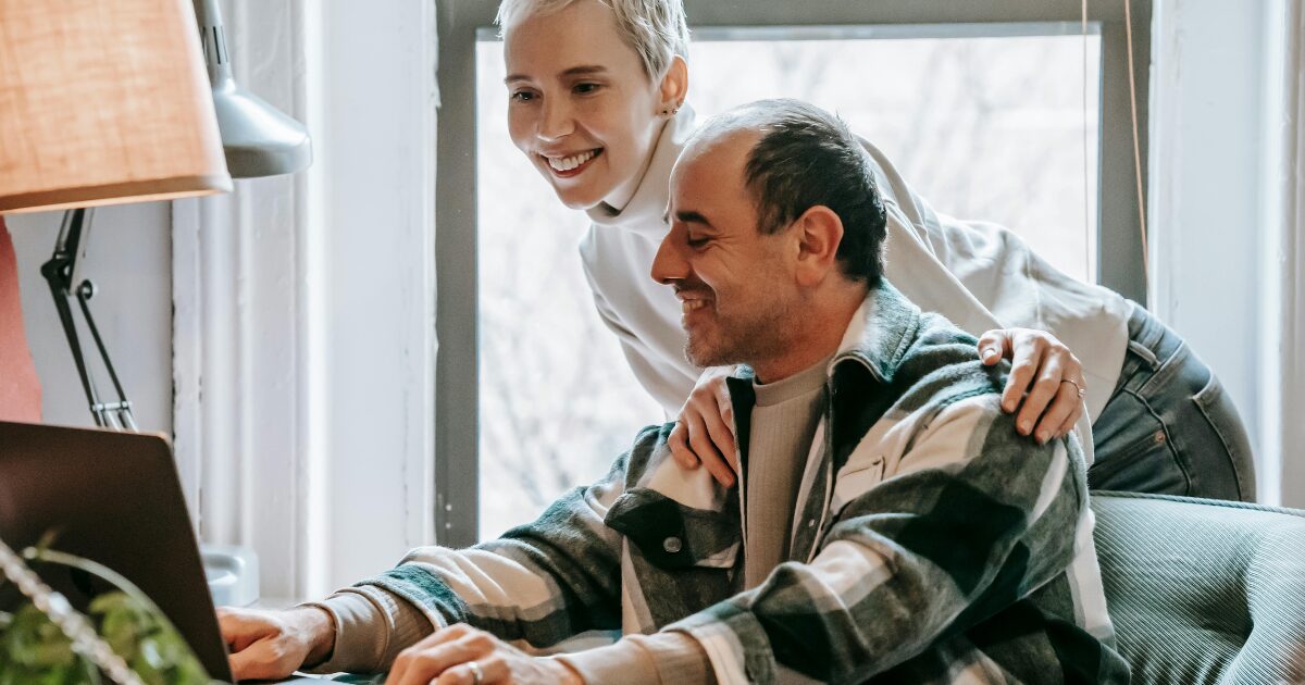 369 A Caucasian man wearing a plaid shirt sits in front of a laptop while a Caucasian woman wearing a cream sweater places her hands on his shoulders.