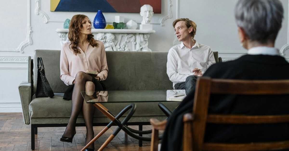 370 A Caucasian woman wearing a pastel pink blouse sits on a grey sofa beside a Caucasian man wearing a white shirt. They are both sitting across from another person shown from behind.
