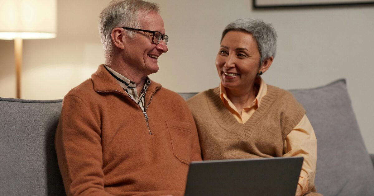 An elderly Caucasian man wearing an orange jacket smiles while sitting beside an elderly Caucasian woman wearing a brown shirt.