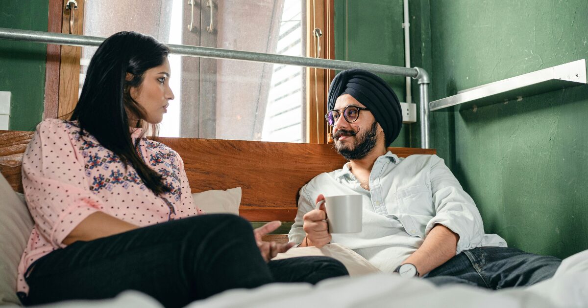 A South Asian man wearing a white shirt looks across at a South Asian woman wearing a pink blouse while holding a cup.