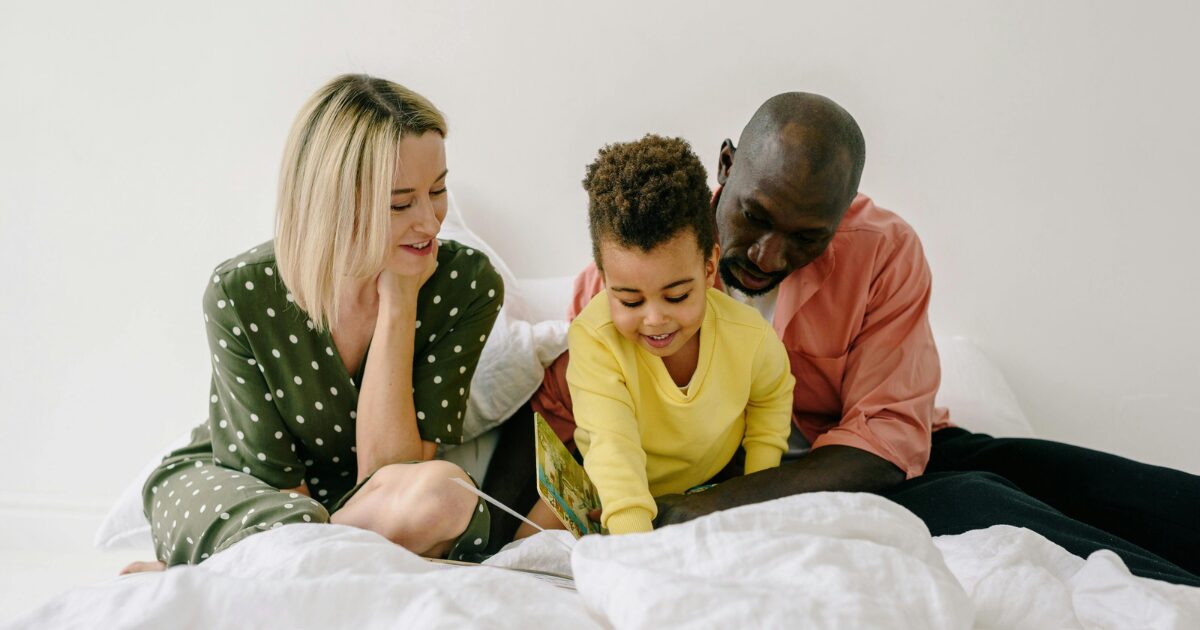 A Caucasian woman wearing a green polka dot blouse and a Black man wearing an orange shirt sit together with a Black child wearing a yellow sweater. They are all reading a children's book.