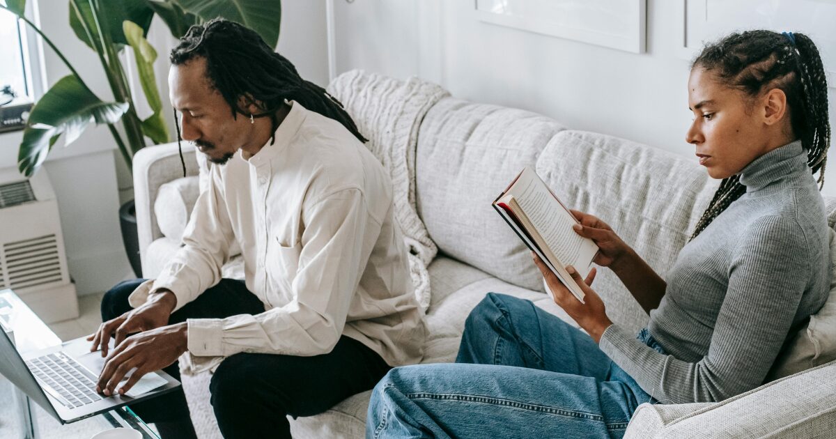 A Black woman wearing a grey sweater sits on a light grey couch reading a book, while a Black man wearing a cream shirt sits beside her on the same couch while using a laptop.