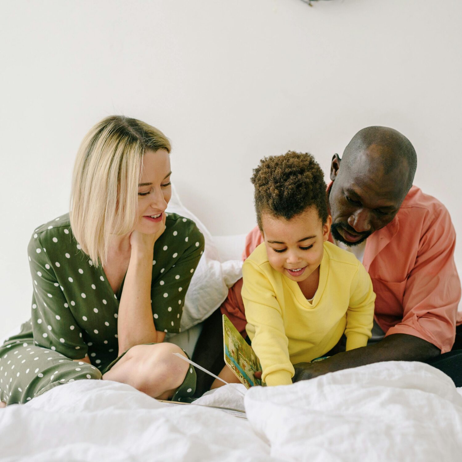 A Caucasian woman wearing a green polka dot blouse and a Black man wearing an orange shirt sit together with a Black child wearing a yellow sweater. They are all reading a children's book.