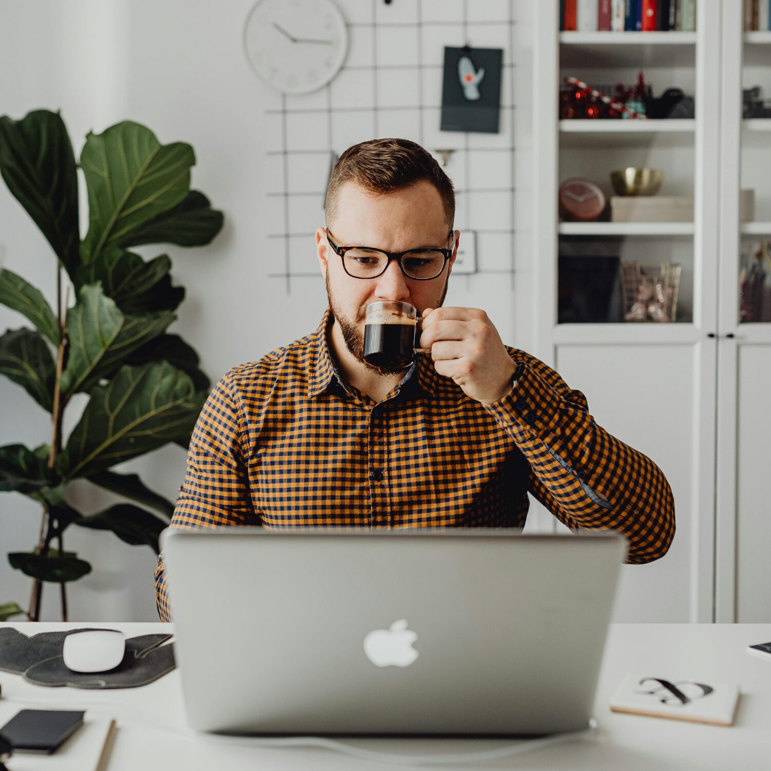 A Caucasian man wearing an orange checkered shirt sipping on a cup while looking at a laptop
