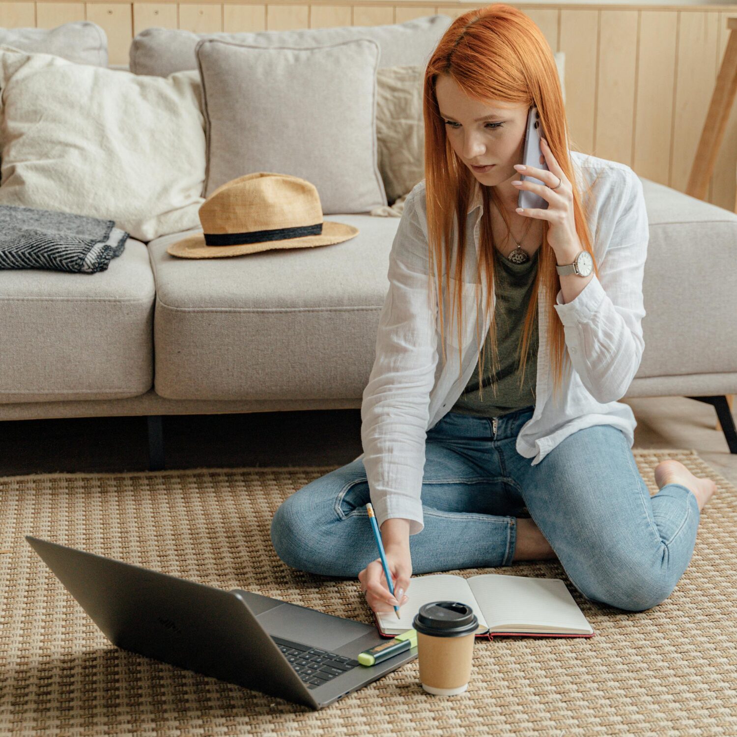 A Caucasian woman wearing a white blazer sits on the floor, writing in a notebook while holding a phone in her left hand.