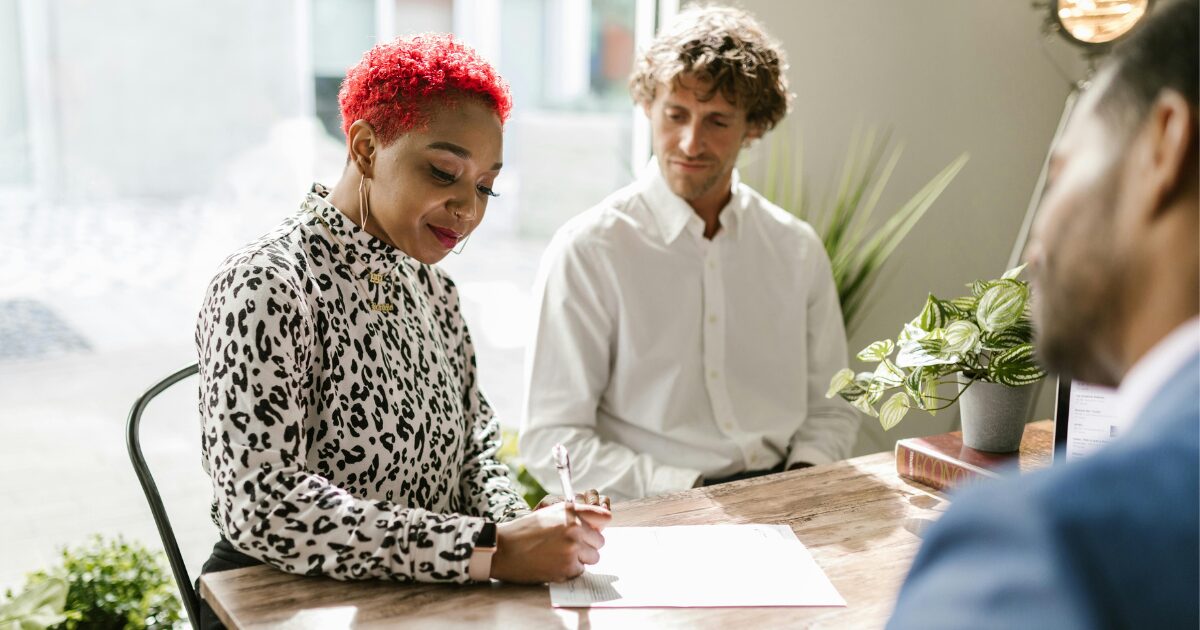 A Black woman wearing an animal-print blouse writes on a document while a Caucasian man wearing a white long-sleeve shirt watches her write..