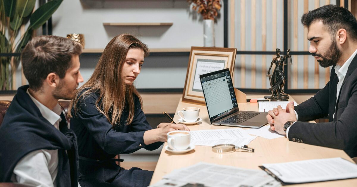 A Caucasian woman wearing a black blouse writes on a document while one Caucasian man wearing a black business suit and another Caucasian man wearing a white long-sleeve shirt watch her write.