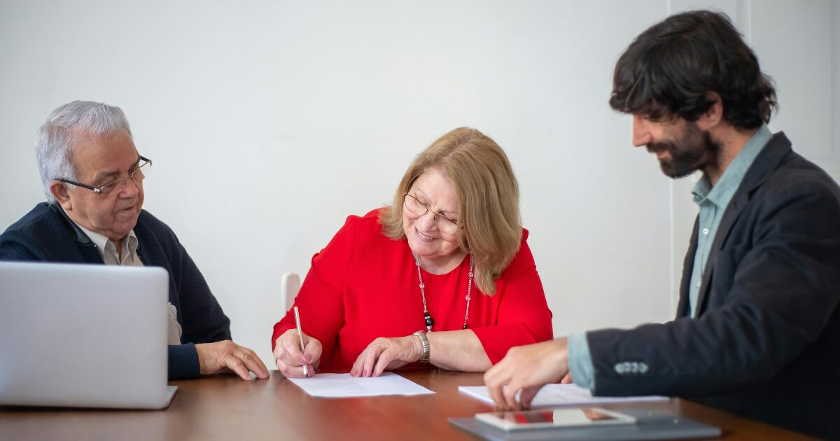 A Caucasian woman wearing a red blouse signs a document as two Caucasian men wearing black business attire watch her.