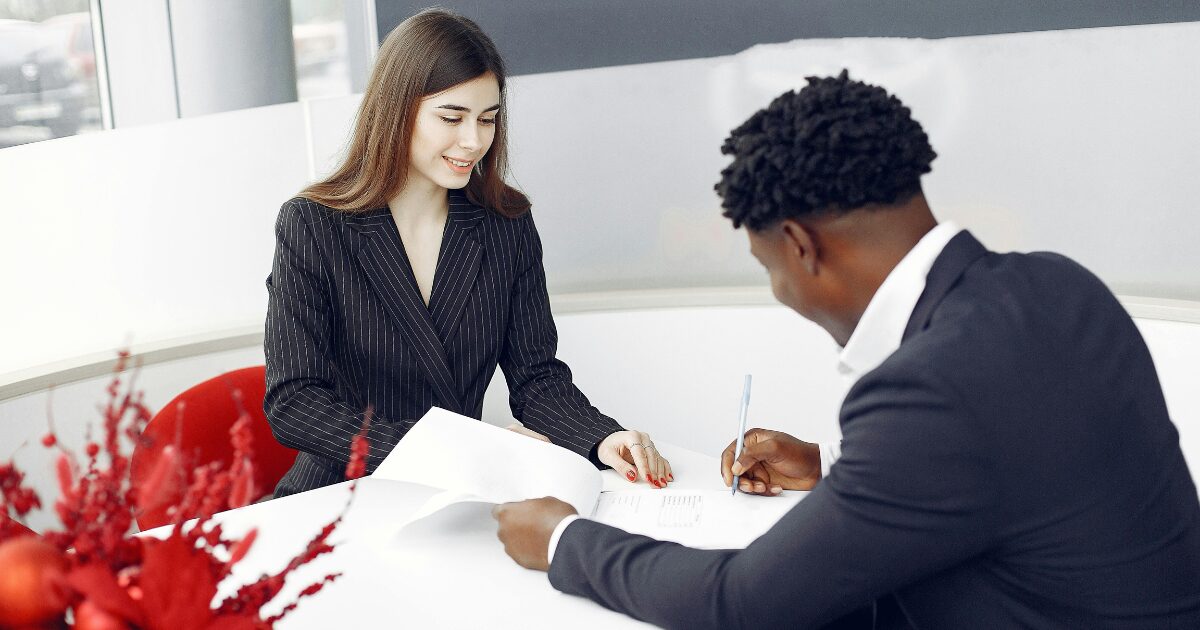 A Black man wearing a black business suit writes on a document while a Caucasian woman wearing a black blazer watches.