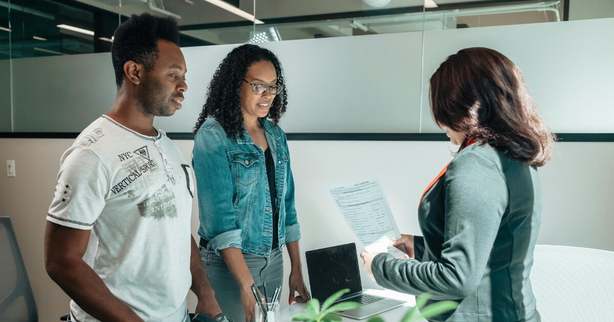 A Black woman wearing a denim jacket and a Black man wearing a white shirt stand in front of a woman holding a document