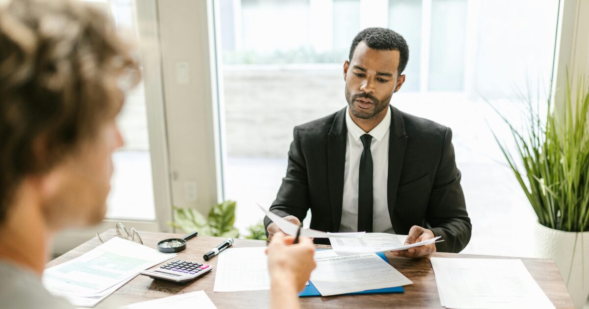 A Black man wearing a black business suit hands a document to a man seated across from him.