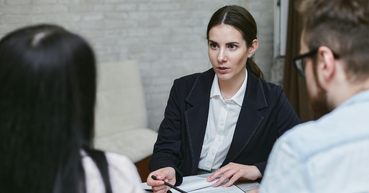 A Caucasian woman wearing a black blazer is speaking with a man and a woman while pointing a pen toward a document with her right hand.