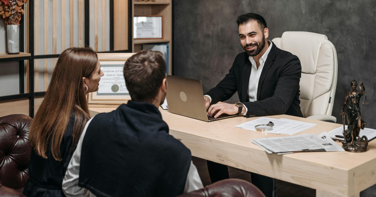 A Caucasian man wearing a black blazer smiles at a man and a woman seated across from him while using a laptop.