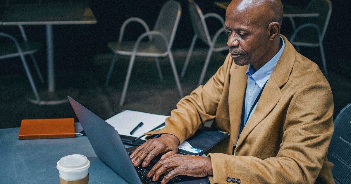 A Black man wearing a mustard-colored blazer is typing on a laptop.