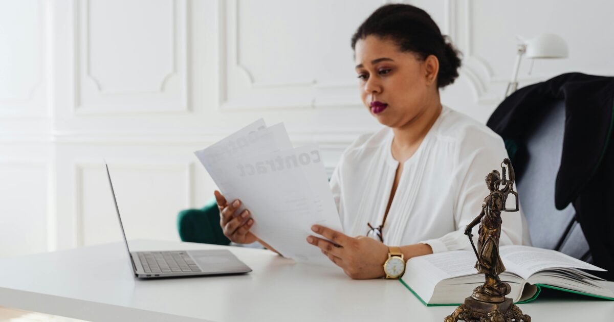 A Black woman wearing a white long-sleeve blouse is reading documents with an open laptop in front of her.
