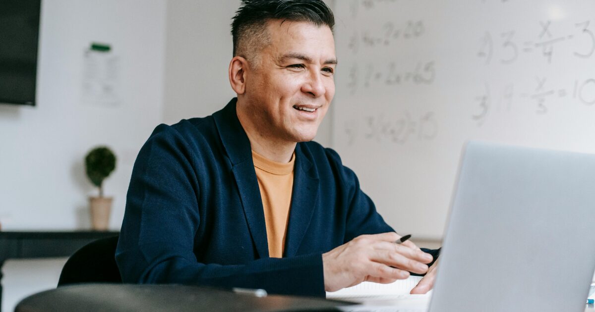 A Caucasian man wearing a navy blue blazer smiles in front of a laptop.