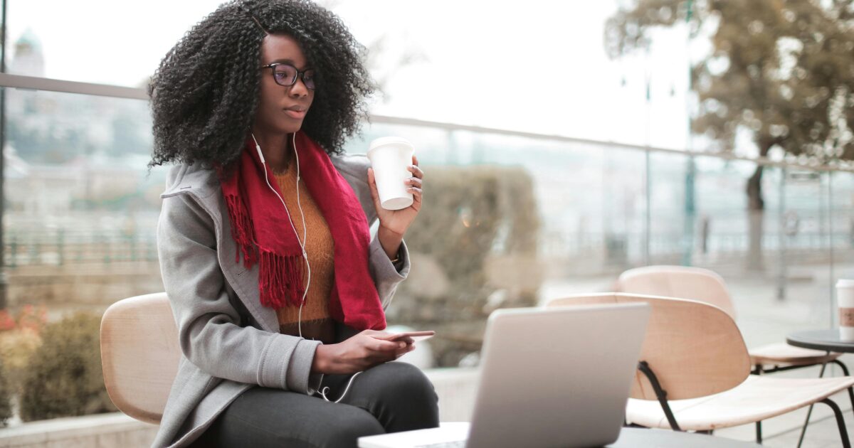 A Black woman wearing a grey blazer and a red scarf, sitting with a cup of coffee in hand while looking at her laptop.
