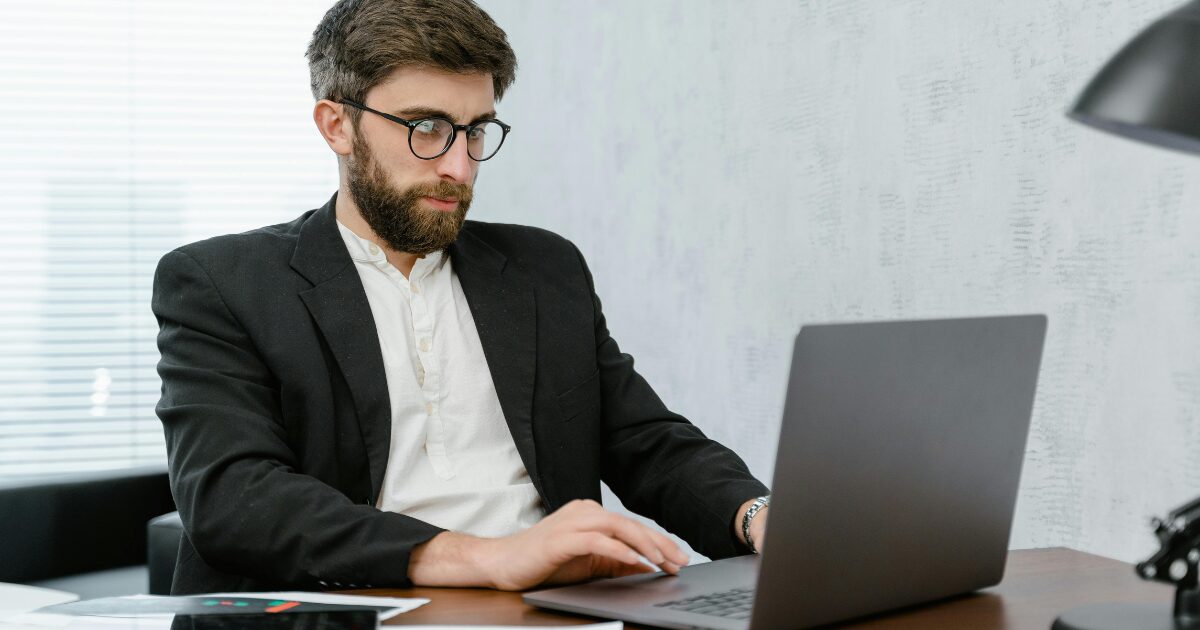 A Caucasian man wearing a black blazer sits while using his laptop.