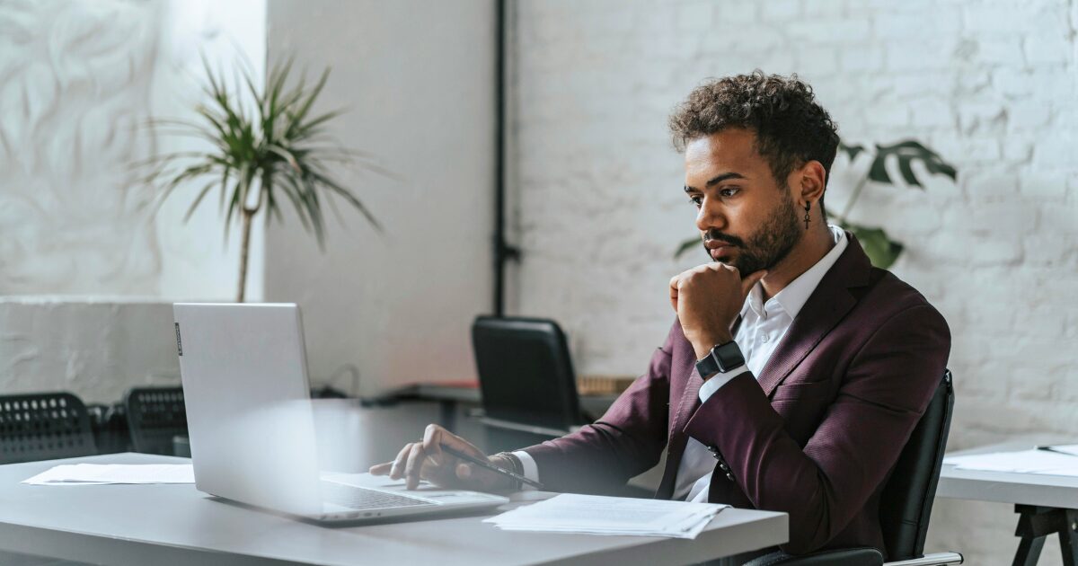 A South Asian man wearing a maroon business suit sitting while using his laptop.