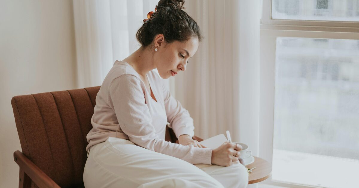A Caucasian woman wearing a light pink blouse sits on a chair, writing something down.