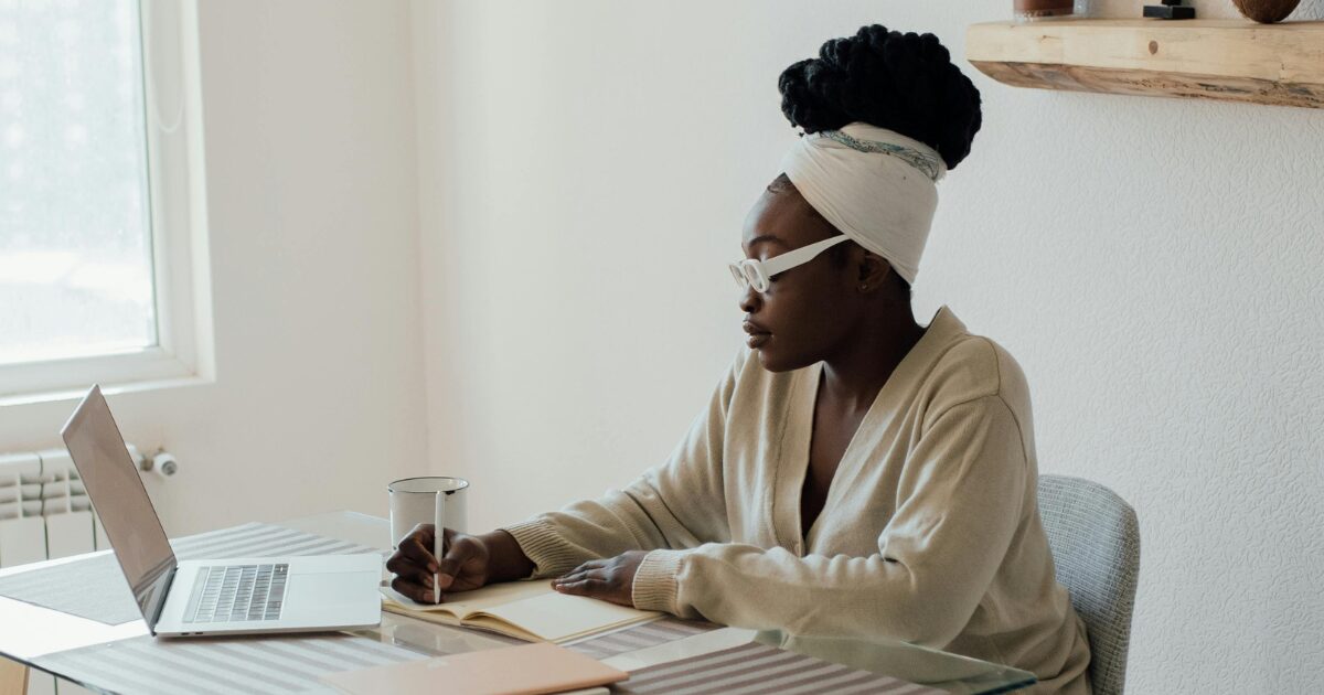 A Black woman wearing a cream-coloured sweater sits on a chair, writing in a notebook.
