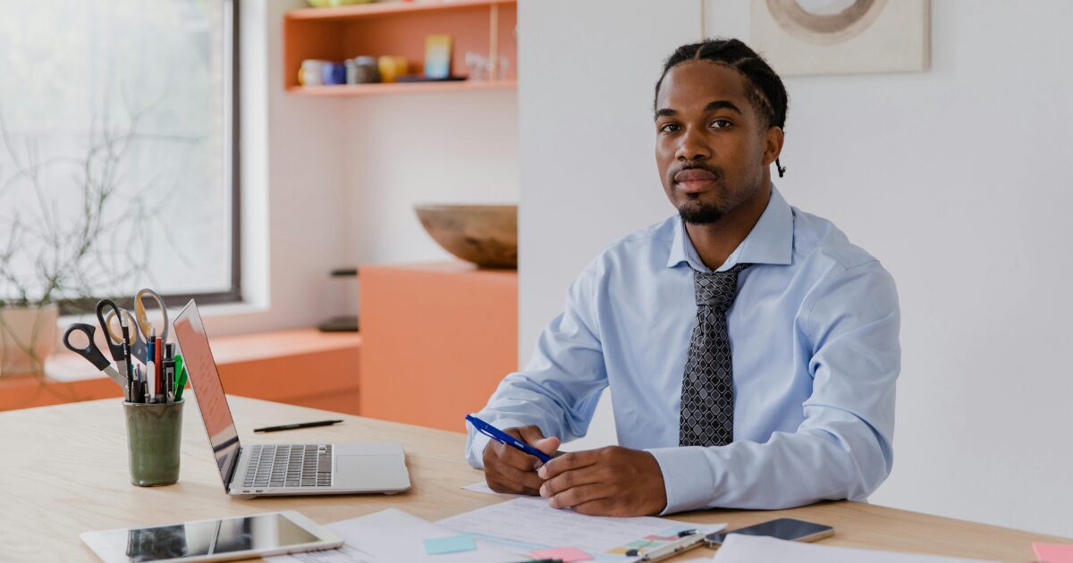 A Black man wearing a light blue shirt and a grey tie sits in front of an open laptop, holding a pen in his hands.