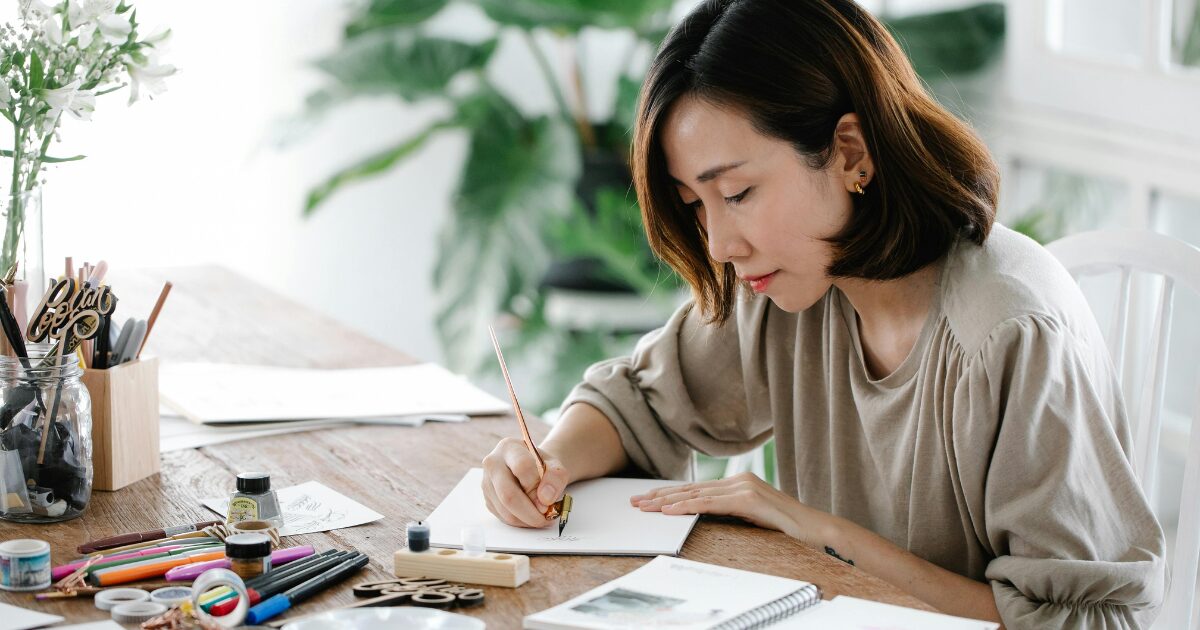 An East Asian woman wearing a light brown blouse writes on a piece of paper.
