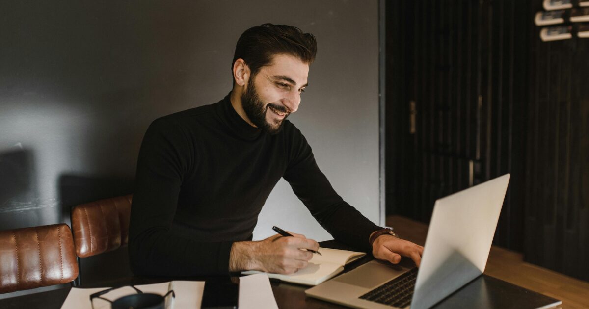 A Caucasian man wearing a black long-sleeve shirt smiles as he holds a pen in his right hand and looks at his laptop.