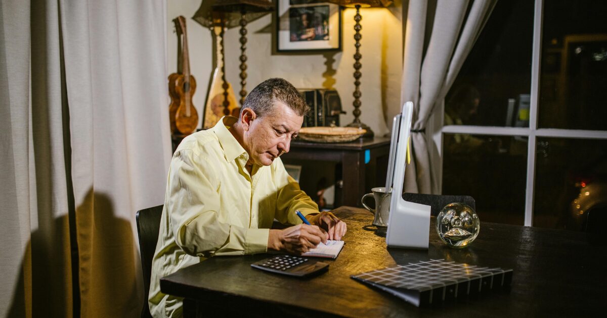 An elderly Caucasian man wearing a yellow long-sleeved shirt sits while writing in a notebook.