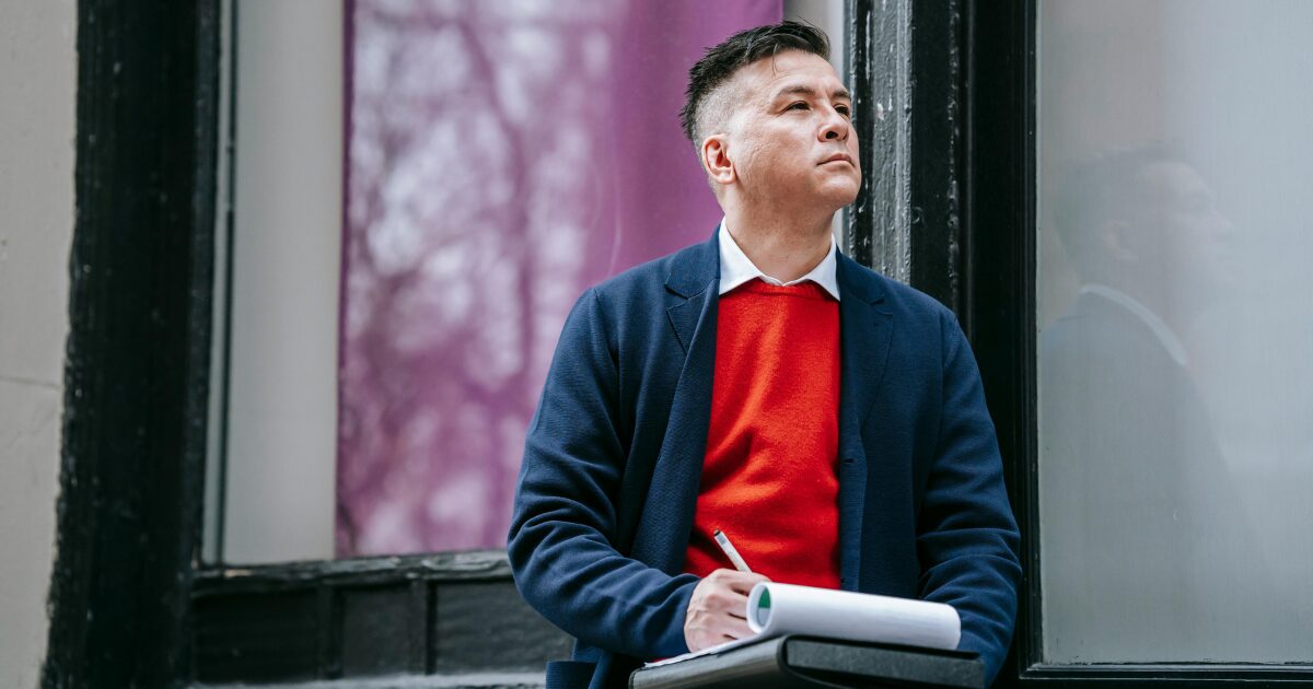 A Caucasian man wearing a navy blue blazer looks up while holding a pen and documents on his lap.