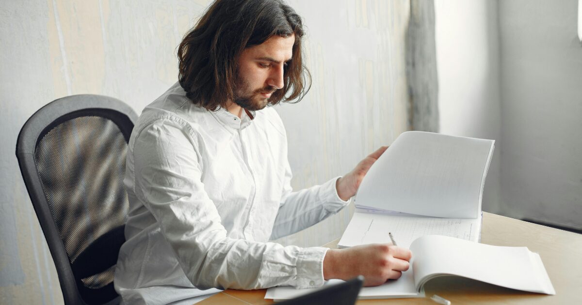 A Caucasian man wearing a white long-sleeve shirt writes on a document as he flips through pages with his left hand.