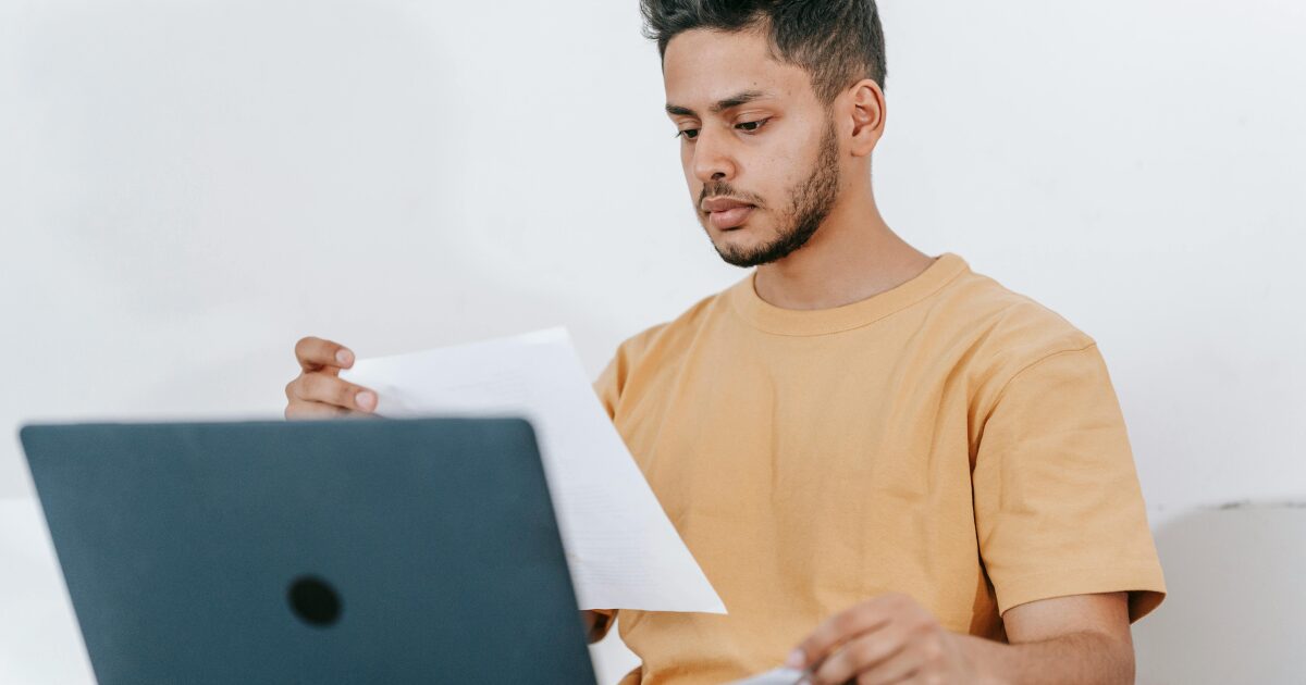 A South Asian man wearing a light orange t-shirt reads a document while sitting in front of an open laptop