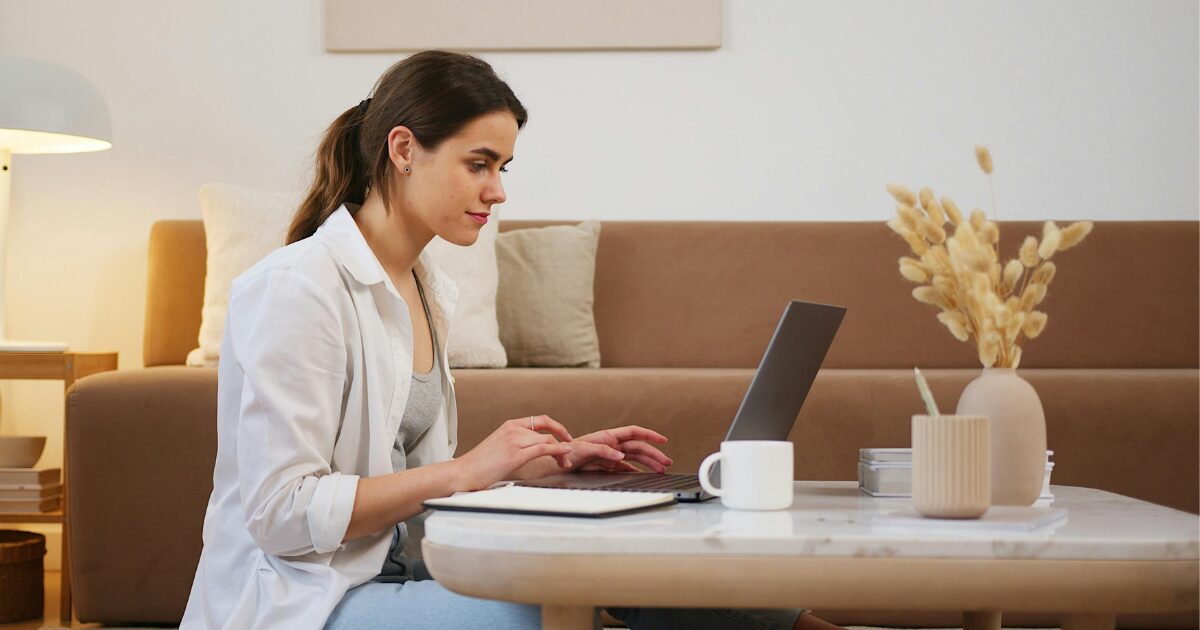 A Caucasian woman wearing a white blouse, sitting on the floor and using her laptop.