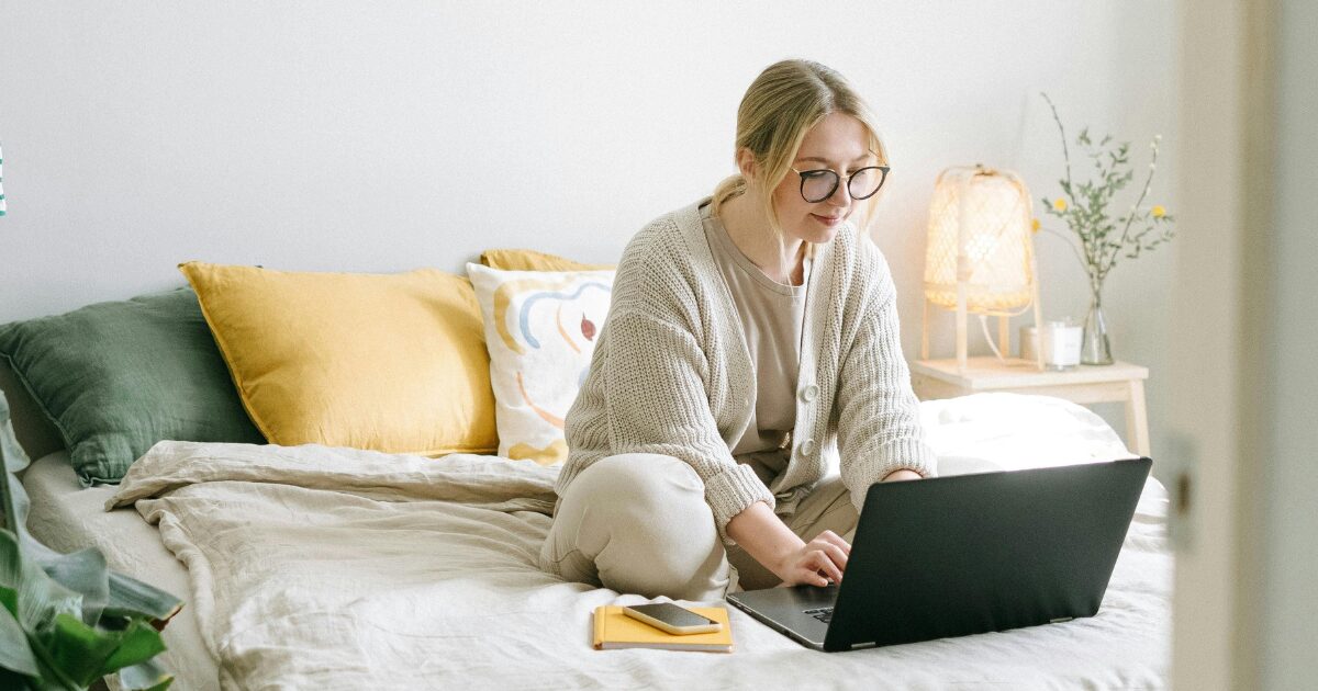 A Caucasian woman wearing a cream-coloured cardigan sits cross-legged on her bed while using a laptop.