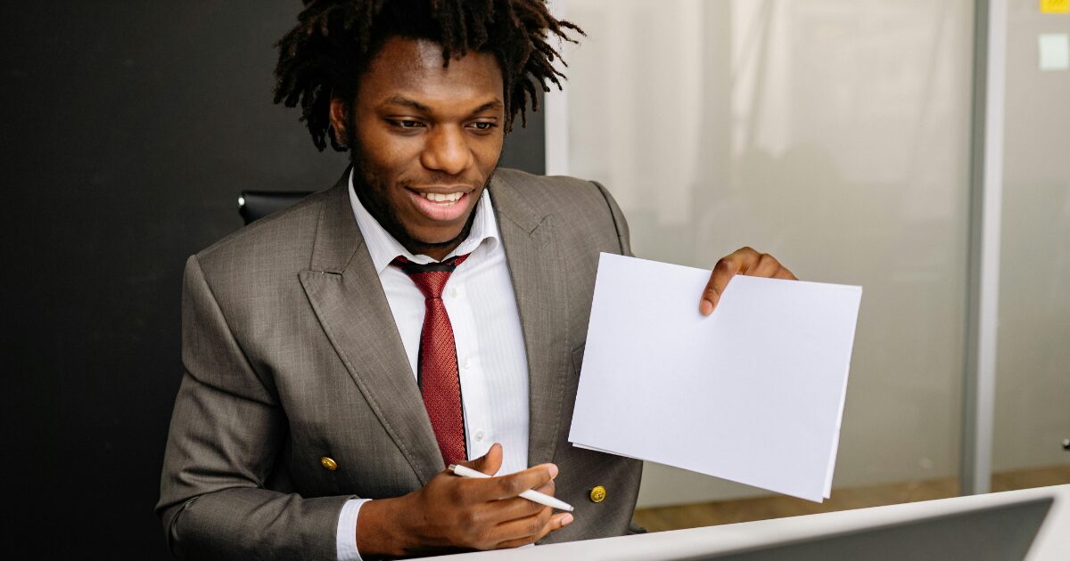 A Black man wearing a grey business suit, holding documents in his left hand and a pen in his right hand.