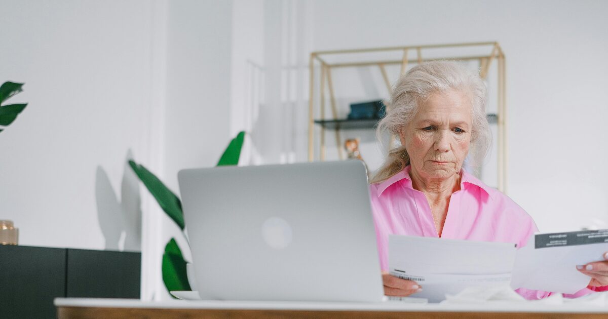An elderly Caucasian woman wearing a pink blouse, holding documents in her hand while sitting in front of an open laptop.