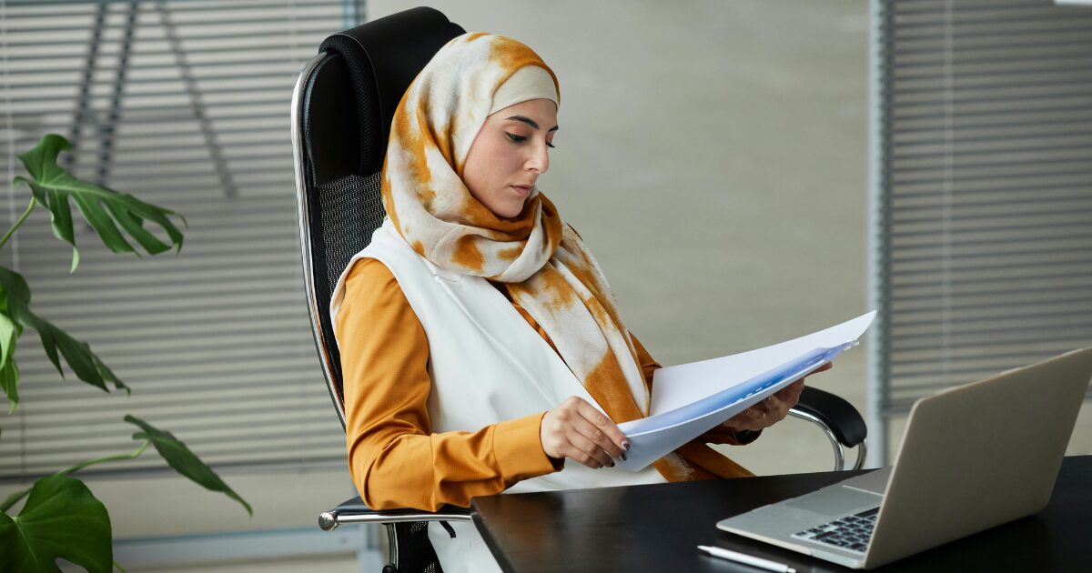 A Middle Eastern woman wearing an orange long-sleeve blouse, reading documents while sitting in front of an open laptop.